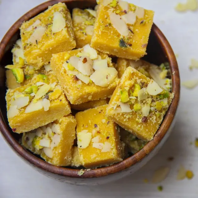 Squares of yellow barfi with sliced almonds in a bowl